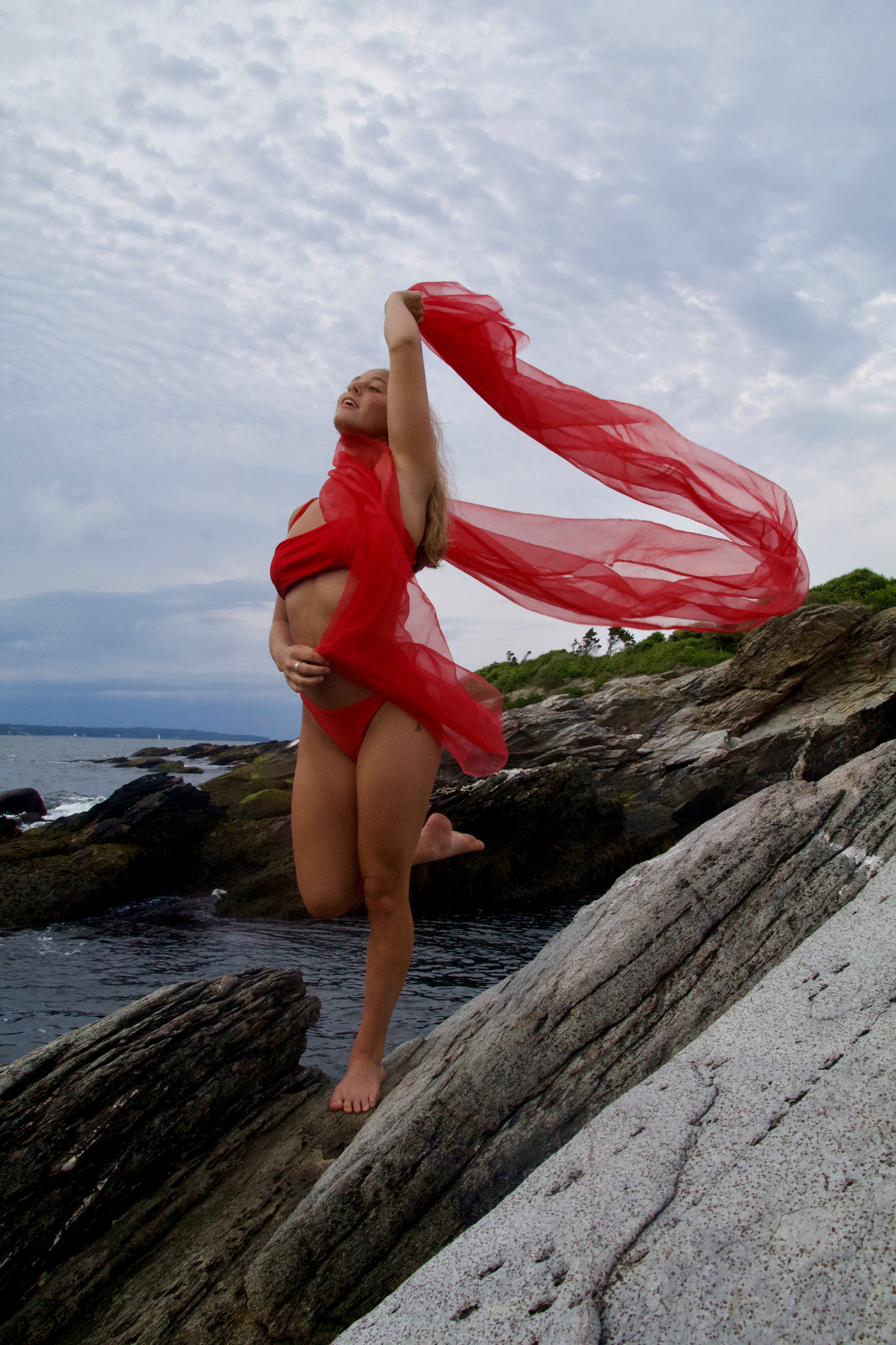 A lady draped in red chiffon standing on a rock by the sea. 